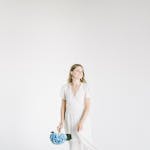 Elegant woman in white dress holding a blue hydrangea bouquet against a minimalist background.